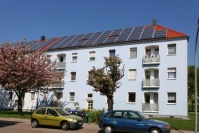 Gebäudezeile mit drei Wohnetagen und hellblauer Fassade mit Loggien, davor blühende Bäume. Solarpanels auf Dach.