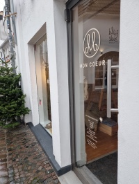 Schräger Blick auf die Eingangstür des Mon Coeur-Pop-up-Stores mit Schriftzug und Logo des Betriebs, am Rand link steht ein Tannenbaum