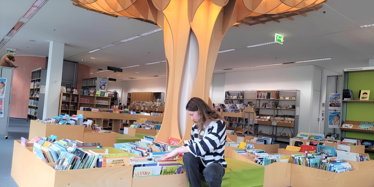 Eine Frau mit langen Haaren sitzt vor einem Bücherregal in einer Bibliothek, umgeben von bunten Kinderbüchern. Über ihr schwebt eine kunstvolle Baumstruktur.