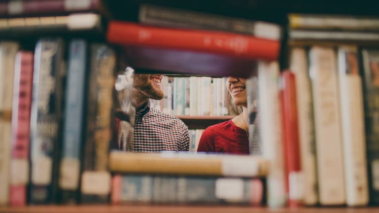 Zwei Personen lächeln sich durch eine Lücke zwischen den Büchern in einem Bibliotheksregal an. Zwei Personen lächeln sich durch eine Lücke zwischen den Büchern in einem Bibliotheksregal an.