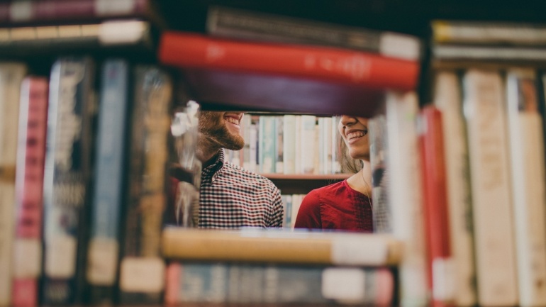 Zwei Personen lächeln sich durch eine Lücke zwischen den Büchern in einem Bibliotheksregal an. Zwei Personen lächeln sich durch eine Lücke zwischen den Büchern in einem Bibliotheksregal an.