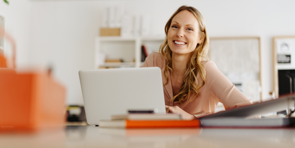 Überweisungen und Daueraufträge Eine lachende blonde Frau sitzt an einem Schreibtisch vor einem aufgeschlagenen Laptop. Foto: contrastwerkstatt / AdobeStock