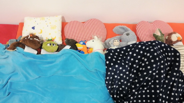 Verschiedene Kuscheltiere liegen zugedeckt in der Bibliothek