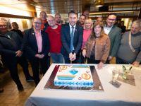 Ein Kuchen wird von einem Mann und einer Frau angeschnitten. Neben Ihnen stehen weitere Menschen und posieren für das Bild