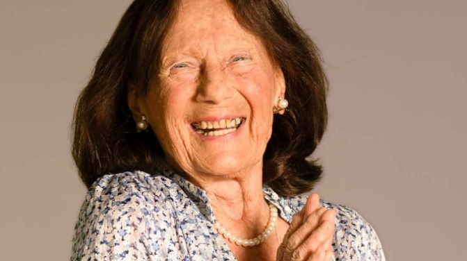 Eine ältere Frau mit langen braunen Haaren lächelt und faltet die Hände. Sie trägt eine geblümte Bluse, eine Perlenkette und Perlenohrringe vor einem einfarbigen Hintergrund.