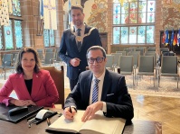 Der Botschafter von Bulgarien, S.E. Grigor Porozhanov, und die Generalsekräterin Diana Popova haben sich bei ihrem Besuch im Saarbrücker Rathaus am 28. Januar in das Goldene Buch der Stadt eingetragen Ein Mann und eine Frau sitzen an einem Tisch in einem Festsaal. Der Mann schreibt etwas in ein Buch. Hinter den beiden steht ein weiterer Mann. Alle drei blicken in die Kamera.