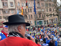 Ein Mann mit schwarzem Hut spricht in ein Mikrofon, während er auf die versammelte Menge auf dem Rathausplatz schaut.