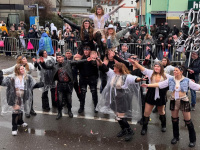 ine Gruppe kostümierter Tänzerinnen und Tänzer führt auf der Straße eine Hebefigur vor, während Zuschauerinnen und Zuschauer hinter Absperrgittern stehen.