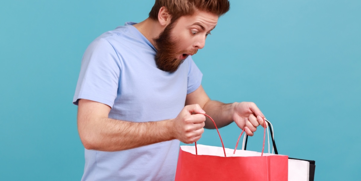 Ein junger Mann mit Bart und in lila T-Shirt steht vor einer blauen Wand und schaut überrascht in eine rote Papiertüte.