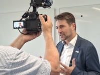 Ein Mann im Anzug gibt vor einer Kamera ein Interview und gestikuliert dabei mit den Händen.