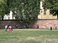 Mehrere Personen sammeln mit Greifzangen Abfall auf einer Grünfläche und entlang einer Mauer und nutzen rote Müllsäcke.
