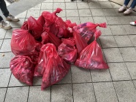 Mehrere gefüllte und zugebundene rote Müllsäcke liegen auf einem gepflasterten Boden. Rundherum sind Beine von Personen zu sehen.