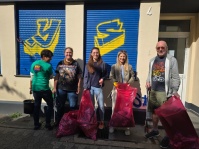Fünf Personen stehen mit Greifzangen und roten Müllsäcken vor einem Gebäude mit blau-gelb bemalten Rollläden.