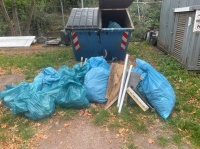Blaue Müllsäcke, Bretter und Metallteile liegen vor einem geöffneten Abfallcontainer im Freien.