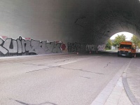 Graffiti an einer Betonwand unter einer Brücke, daneben ein Halteverbotsschild und eine bewachsene Treppe.