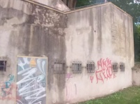 Mit Graffiti besprühte Außenwand eines Gebäudes mit vergitterten Fenstern und einer Tür, davor eine Rasenfläche.