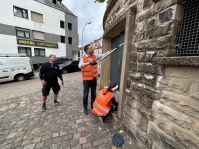 Drei Personen reinigen eine steinerne Gebäudefassade. Zwei von ihnen tragen orangefarbene Warnwesten.