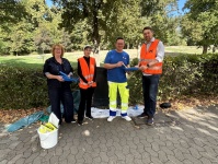 Vier Personen stehen mit Arbeitsmaterialien an einem Weg im Grünen. Zwei tragen orangefarbene Warnwesten.