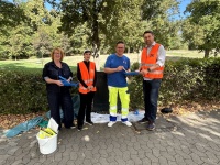 Vier Personen stehen mit Arbeitsmaterialien an einem Weg im Grünen. Zwei tragen orangefarbene Warnwesten.