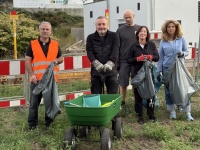 Fünf Personen stehen mit Müllsäcken und Handschuhen auf einer Grünfläche. Vor ihnen steht ein grüner Handwagen.