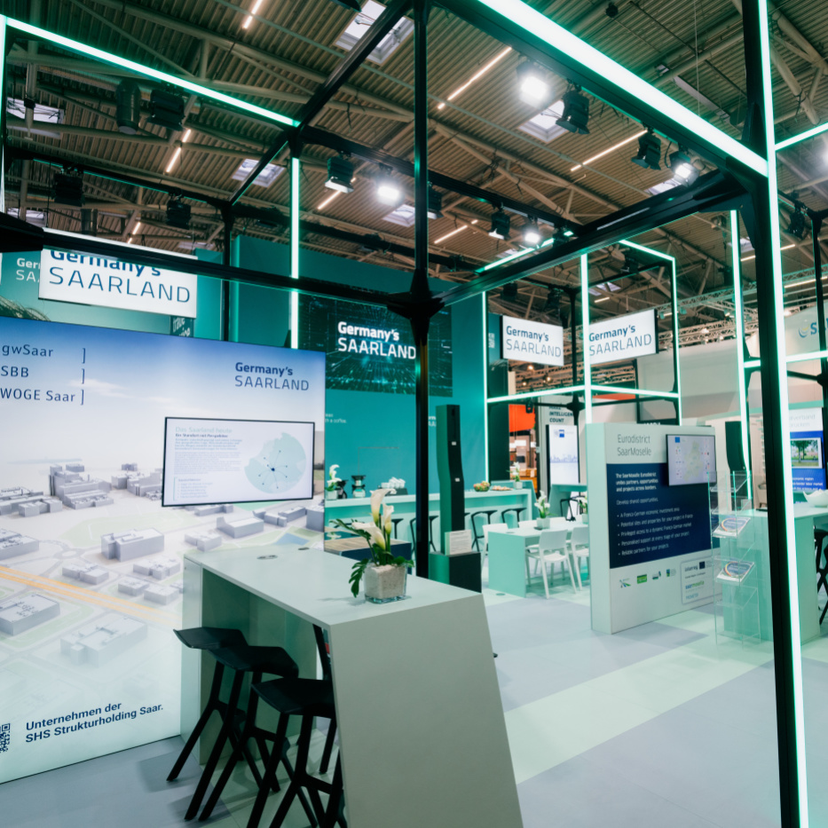 Gemeinschaftsstand "Germany's Saarland" auf der Expo Real 2026 Ein offener mintgrün-leuchtender Messestand mit Hochtischen und -stühlen sowie Messewänden.