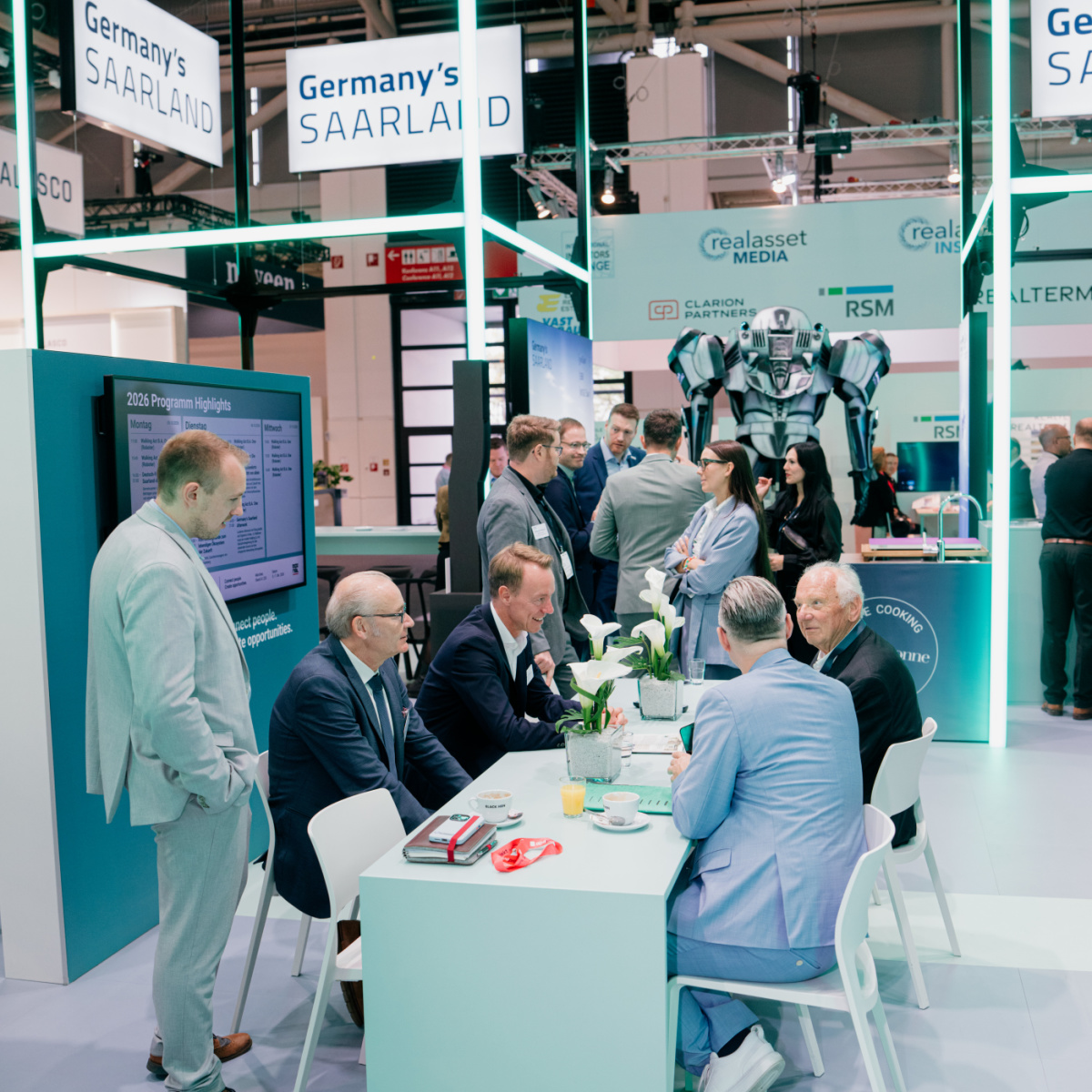 Austausch auf der Expo Real Am Messestand Germany's Saarland sitzen vier Männer an einem Besprechungstisch, ein Mann steht daneben. Im Hintergrund sind weitere Personen, die sich unterhalten.