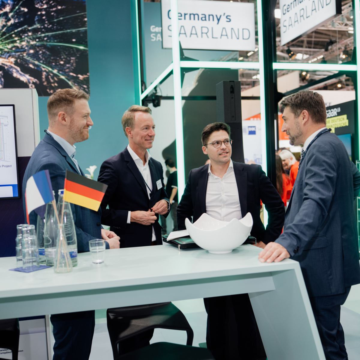 Austausch bei der Expo Real Vier Männer stehen an einem Hochtisch am Messestand und unterhalten sich. Auf dem Tisch steht eine Vase mit einem deutschen und einem französischen Fähnchen.