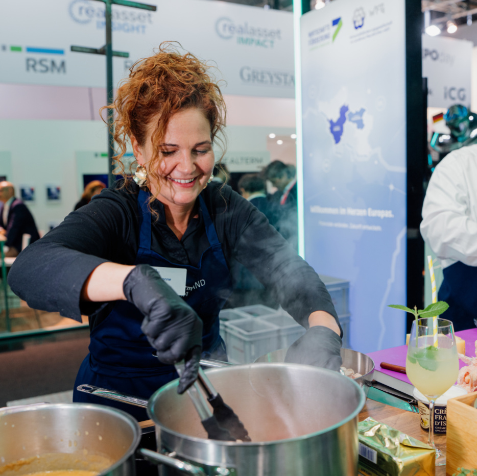 Genuss bei der Expo Real Eine lachende Frau greift mit einer Zange in einen dampfenden Kochtopf. Rechts neben ihr schneidet ein Mann eine Zwiebel mit einem großen Messer.