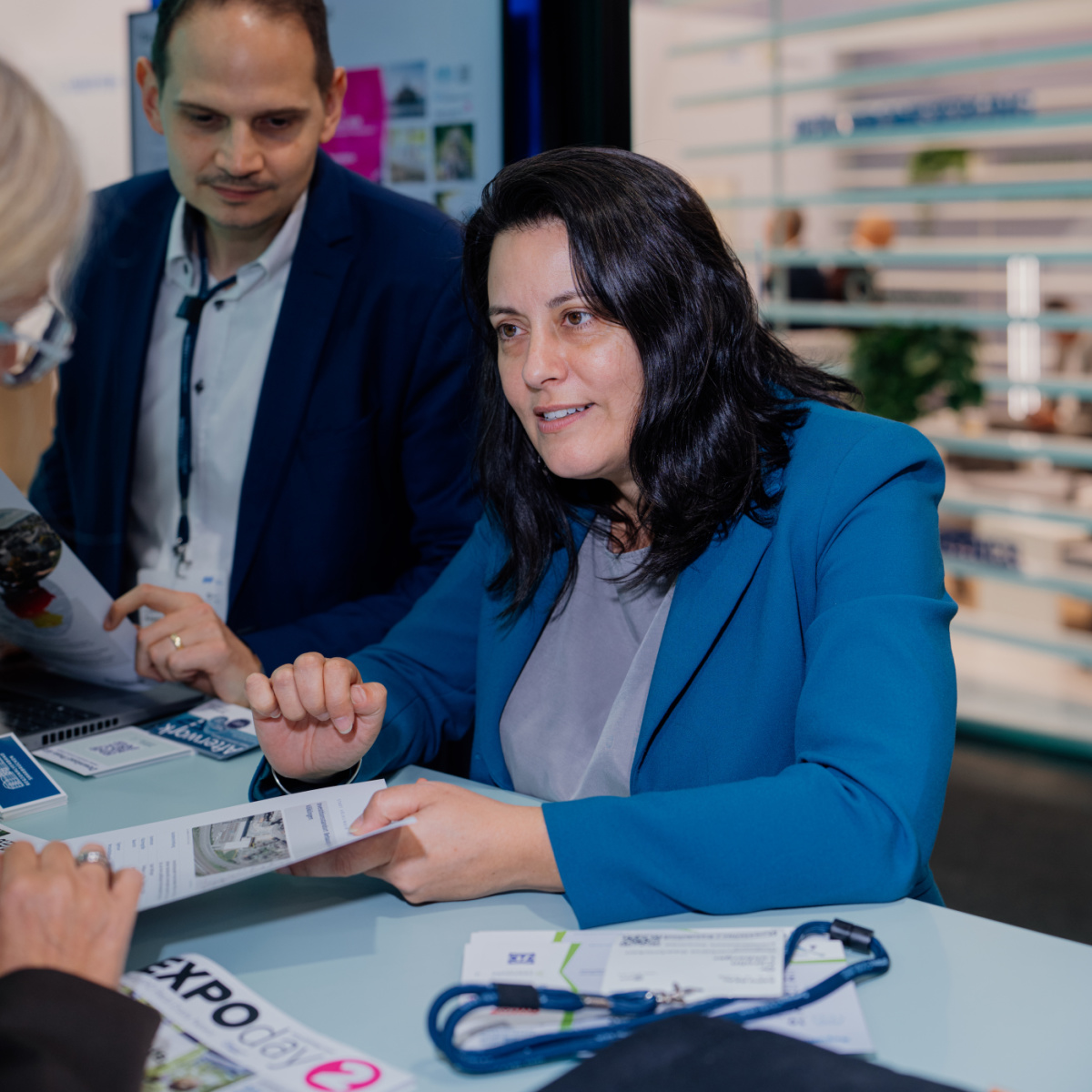 Austausch bei der Expo Real Eine Frau hält ein Blatt in der Hand und unterhält sich mit einer anderen Frau, die gegenüber von ihr am Stand steht.
