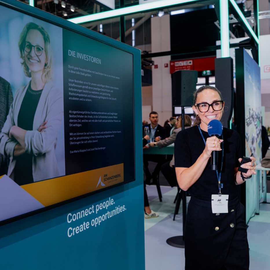 Präsentation am Stand auf der Expo Real Eine Frau mit einem Mikrofon steht vor einer Messewand mit einem Bildschirm, auf dem eine Präsentation abläuft.